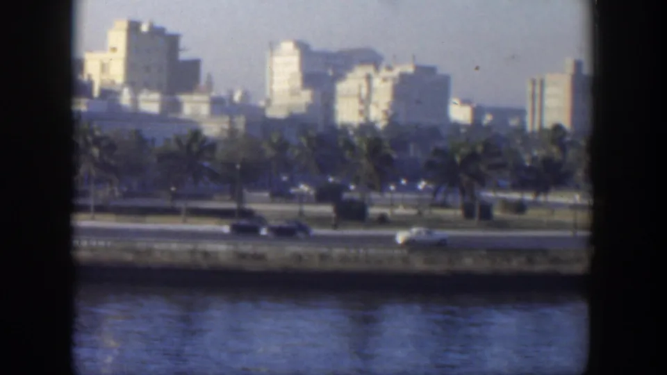 1951 Cuba waterfront road with classic cars passing below palms and a softly focused urban skyline.