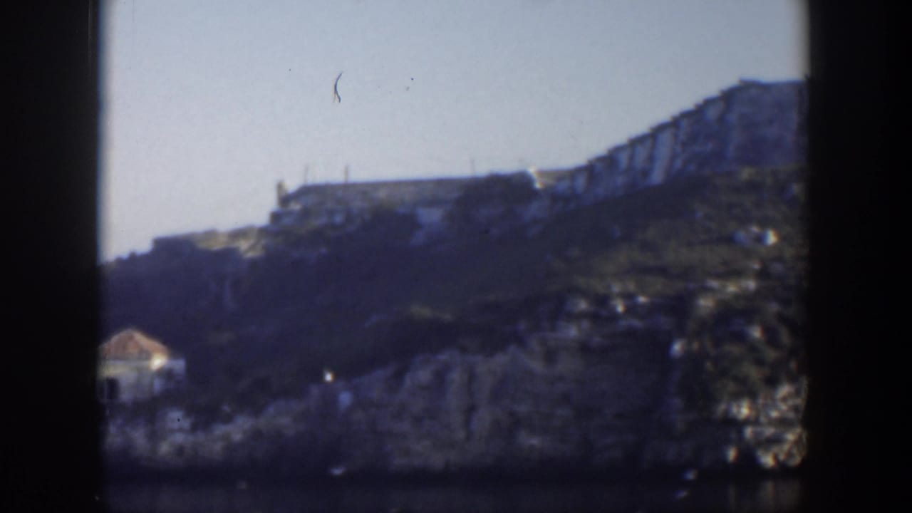 Distant tan building on a rocky green and grey cliff under a pale sky.