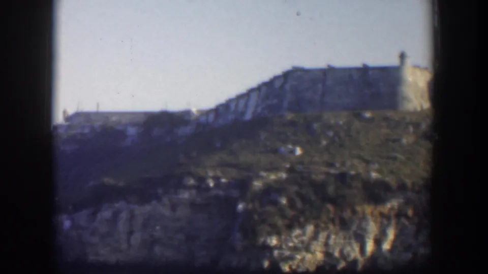 1951 Cuba 8mm view of a tan cliff-edge building, including a tower-like end, above scrub and exposed rock.