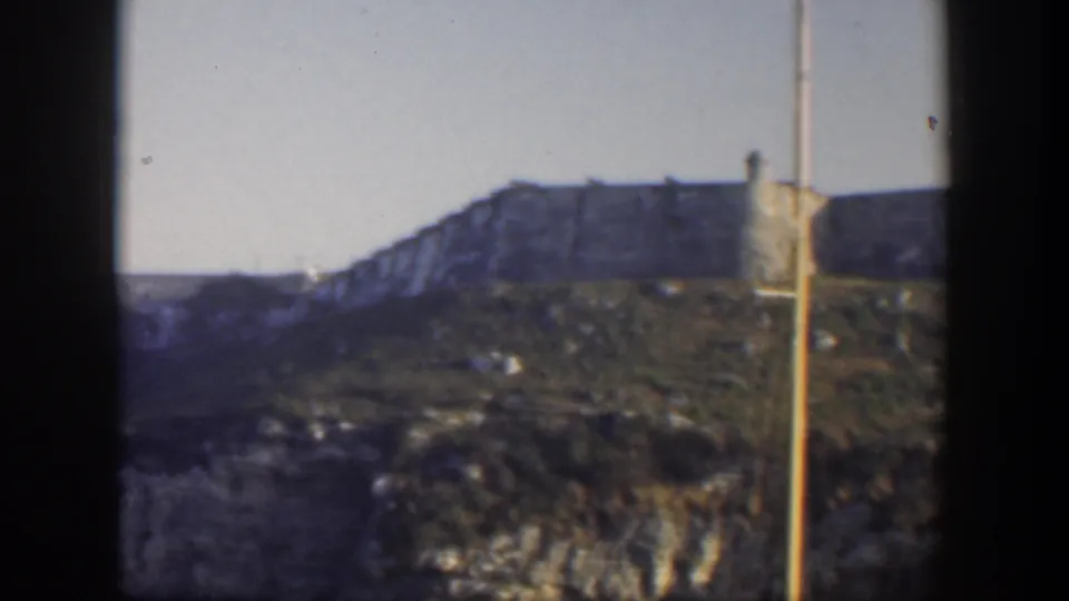 1951 Cuba frame showing the distant tan building above green and grey cliff terrain, with a pale vertical streak near the right edge.