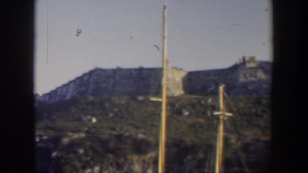 1951 Cuba 8mm view of a weathered stone fortification above a grassy slope, framed by sailboat masts and dark film edges.
