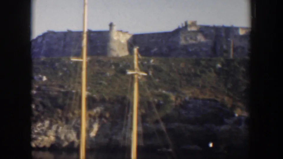 Mid-century Cuba home-movie frame showing weathered stone walls and a small tower on a hillside, partly obscured by sailboat masts.