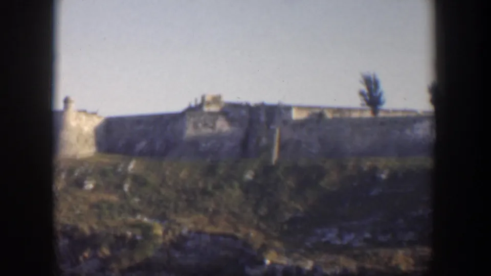 1951 Cuba home-movie view of a stone wall and small tower along a grassy ridge, with a dark tree shape near the right edge.