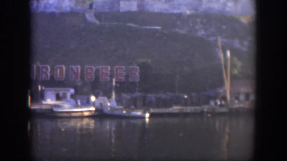 1951 Cuba 8mm waterfront frame with the Ironbeer sign, several small boats, calm water, and a shoreline structure at the right edge.