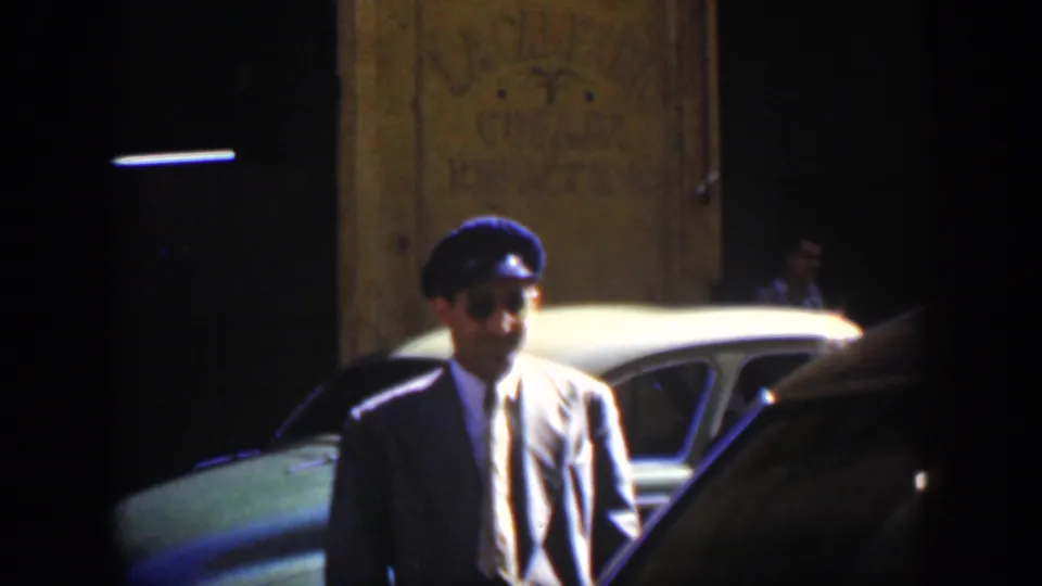 A man in a dark suit, tie, cap, and sunglasses stands near parked vintage cars on a Cuban city street in 1951 footage.