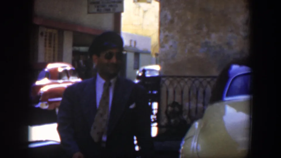 The suited man with cap and sunglasses moves through a sunlit 1951 Cuba street scene beside classic cars.