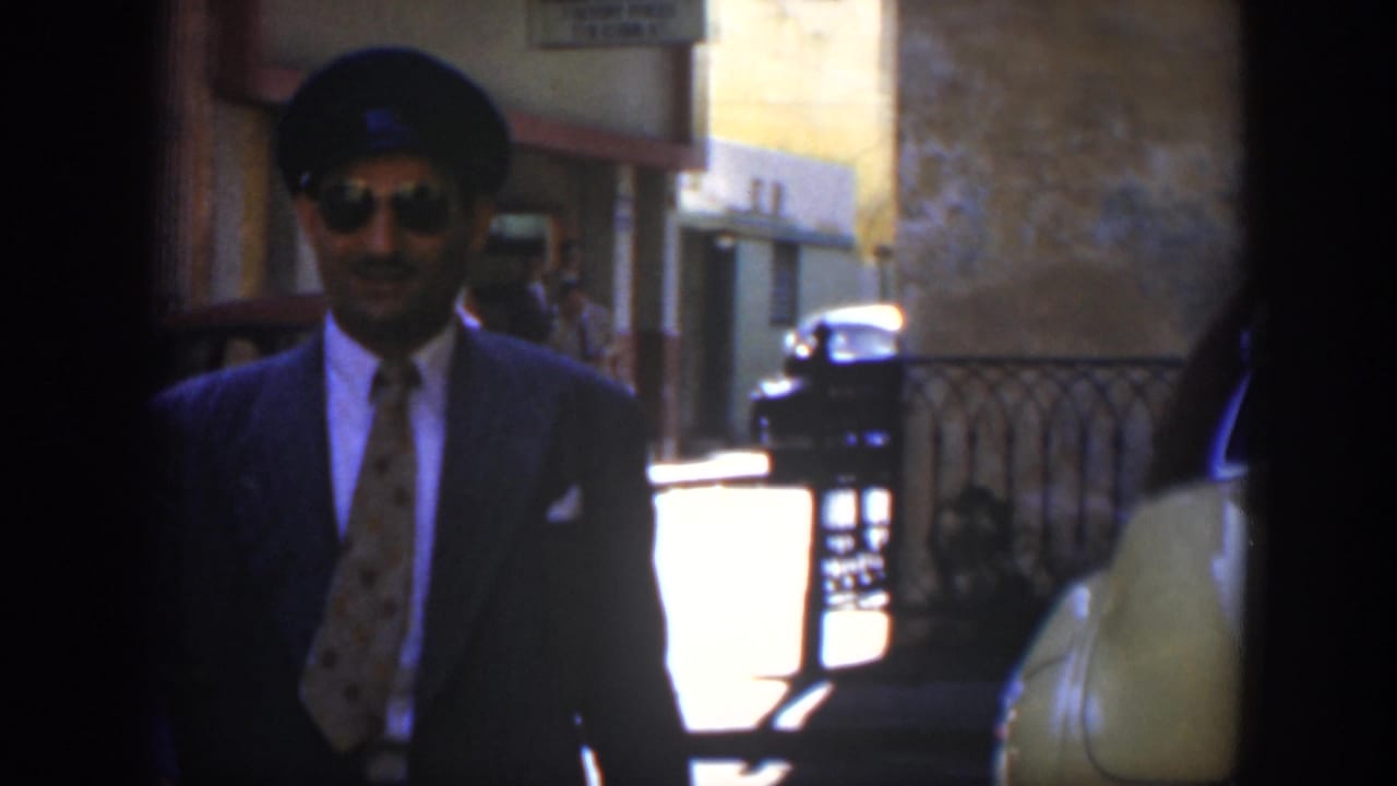 Man in a dark suit and sunglasses walking on a sunlit street near parked cars.