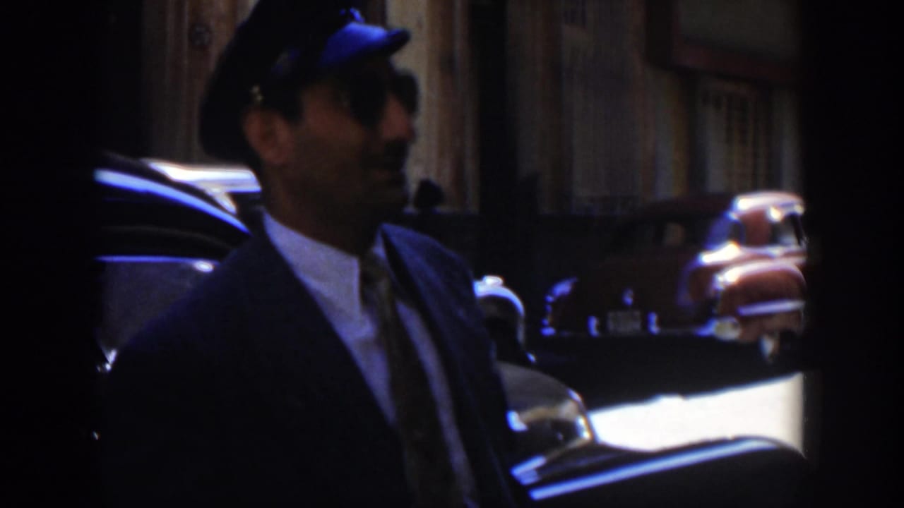 Man in a dark suit and cap seen in profile beside a parked car on a street.