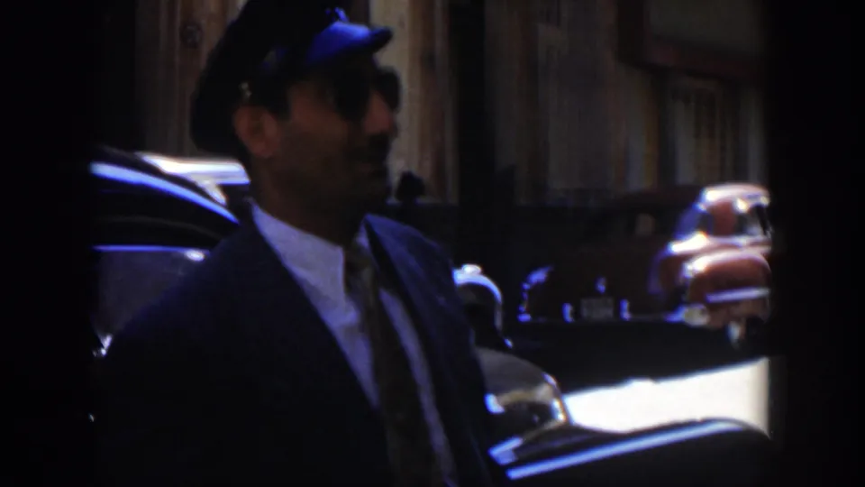 A man in a dark suit and cap stands or walks in profile beside a dark parked car, with older automobiles visible along the street.