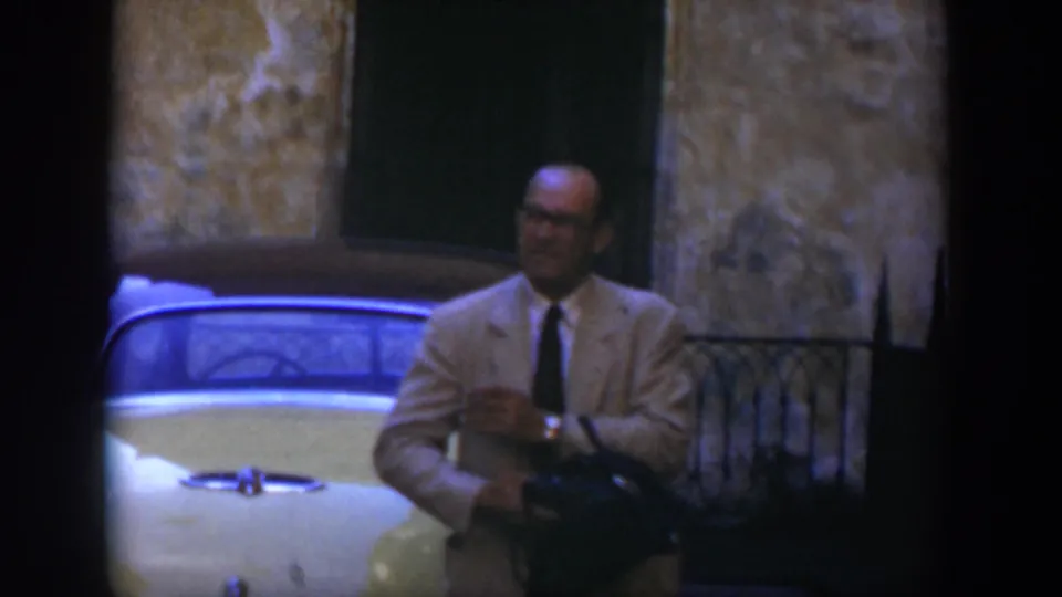 In 1951 Cuba, a man in a light-colored suit and glasses adjusts a dark bag while standing beside a yellow vintage car.