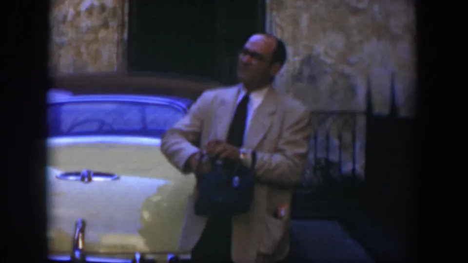 A light-suited man with glasses holds a dark bag near a yellow vintage car, framed by a weathered building wall and dark shutter.