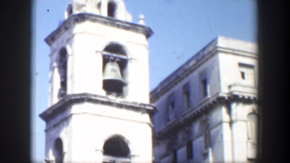 1951 Cuba 8mm frame showing a bright stone bell tower with a large hanging bell, blue sky, and adjacent masonry building.