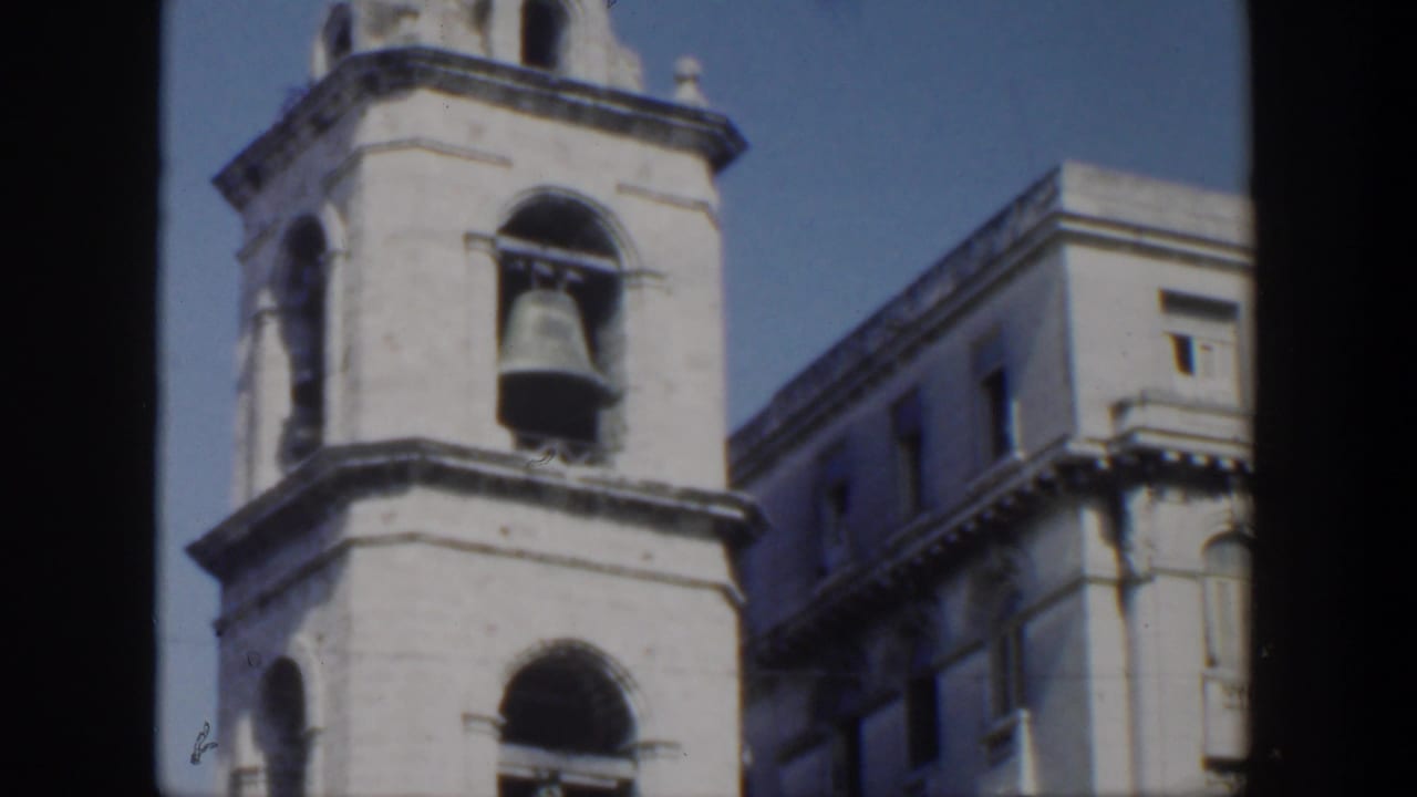 Bell tower with a large bell framed by arched openings and a nearby building.