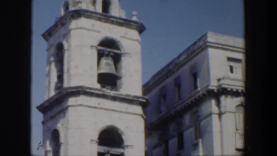 1951 Cuba 8mm frame of a pale masonry bell tower, dark arched openings, blue sky, and an adjoining urban facade.