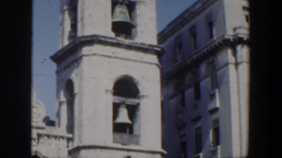 1951 Cuba 8mm frame showing the bell tower more tightly, with arched openings, two bells, and the side of a neighboring building.