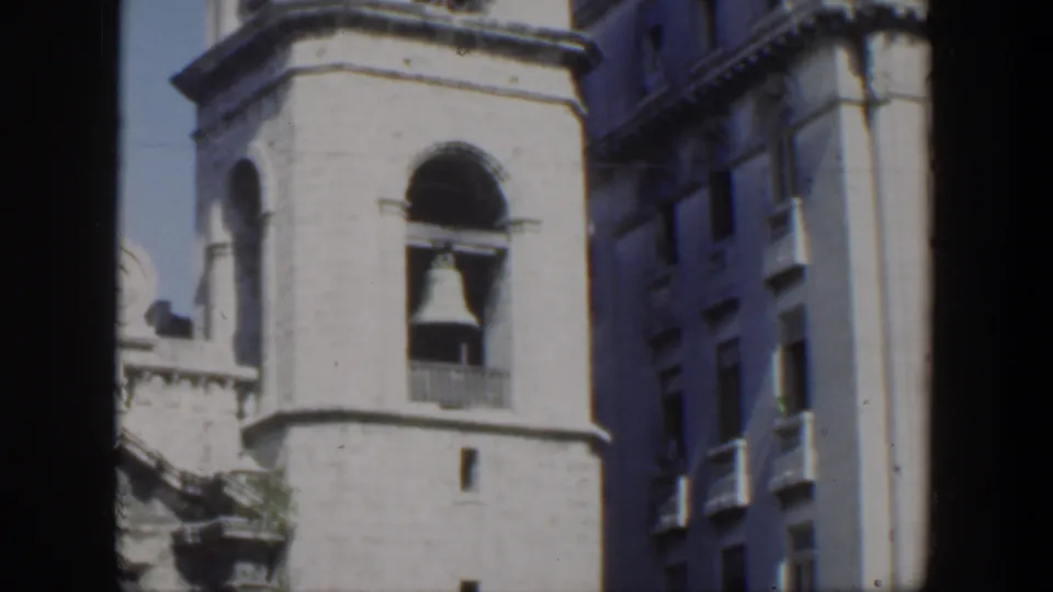 1951 Cuba 8mm frame showing a masonry bell tower with a large bell, arched recesses, blue sky, and neighboring urban architecture.