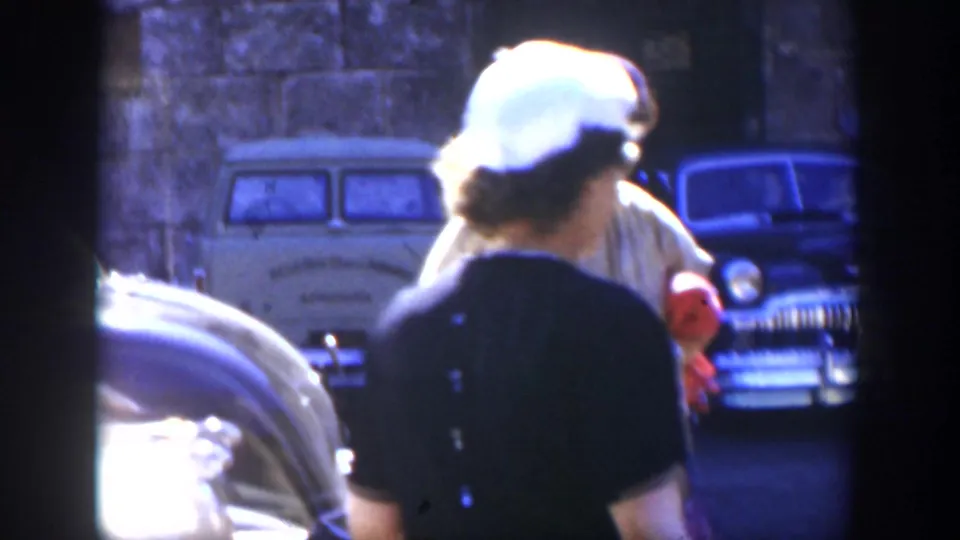 A 1951 Cuba street view shows women in period clothing walking near parked classic automobiles in front of a dark stone wall.