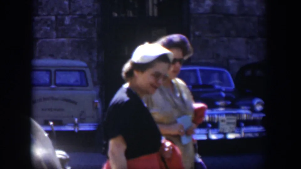 A Cuba 1951 home-movie frame shows two women walking together past parked classic cars, with faces visible and a stone wall behind them.