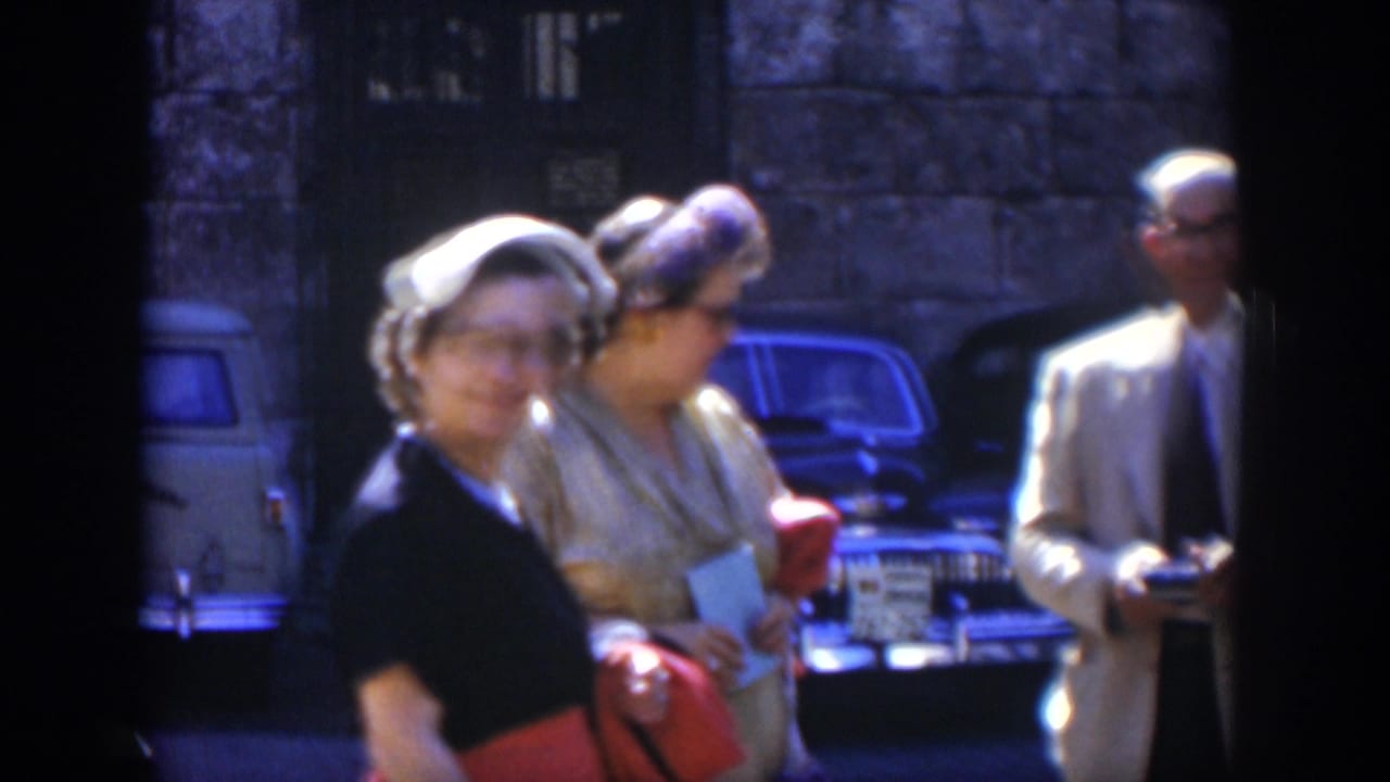 In 1951 Cuba home-movie footage, adults in hats, dresses, and suits stand near parked vintage cars outside a stone building.