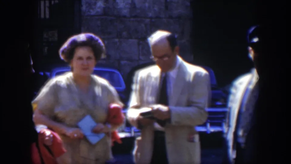 A woman holding papers and a red handbag stands near a man in a light suit, with vintage cars behind them in Cuba.