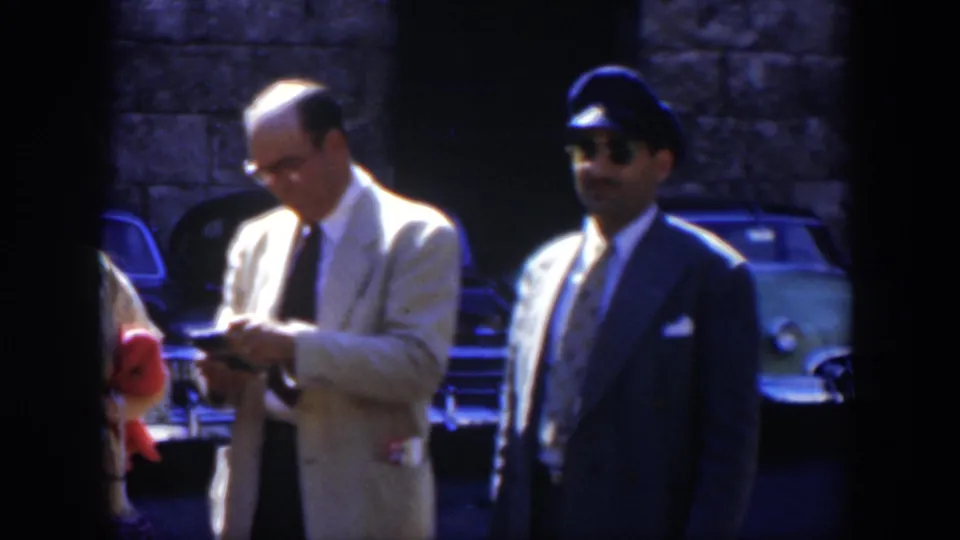 A man in a light suit checks an item as a uniformed man in a cap stands nearby, with vintage cars and stonework behind them.