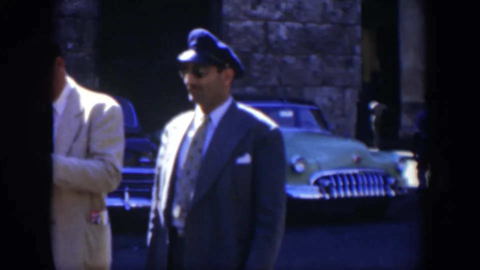 A uniformed man wearing a peaked cap stands in a Cuba parking area, with a vintage car and stone building visible behind him.