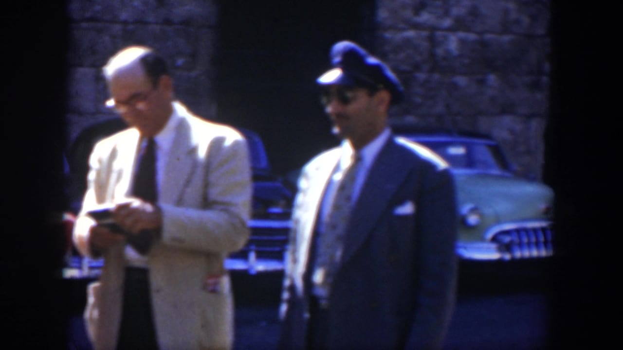 Two men stand on a sunlit street near a classic green car and stone wall.
