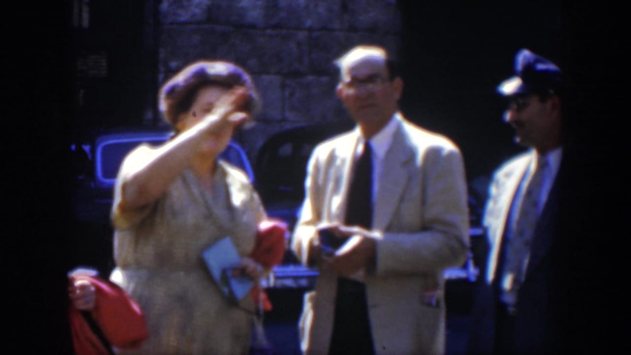 A woman gestures while two suited men stand nearby on a shaded street.
