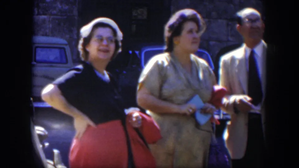 In 1951 Cuba, two women with handbags and a suited man stand near a parked car, looking toward the camera.