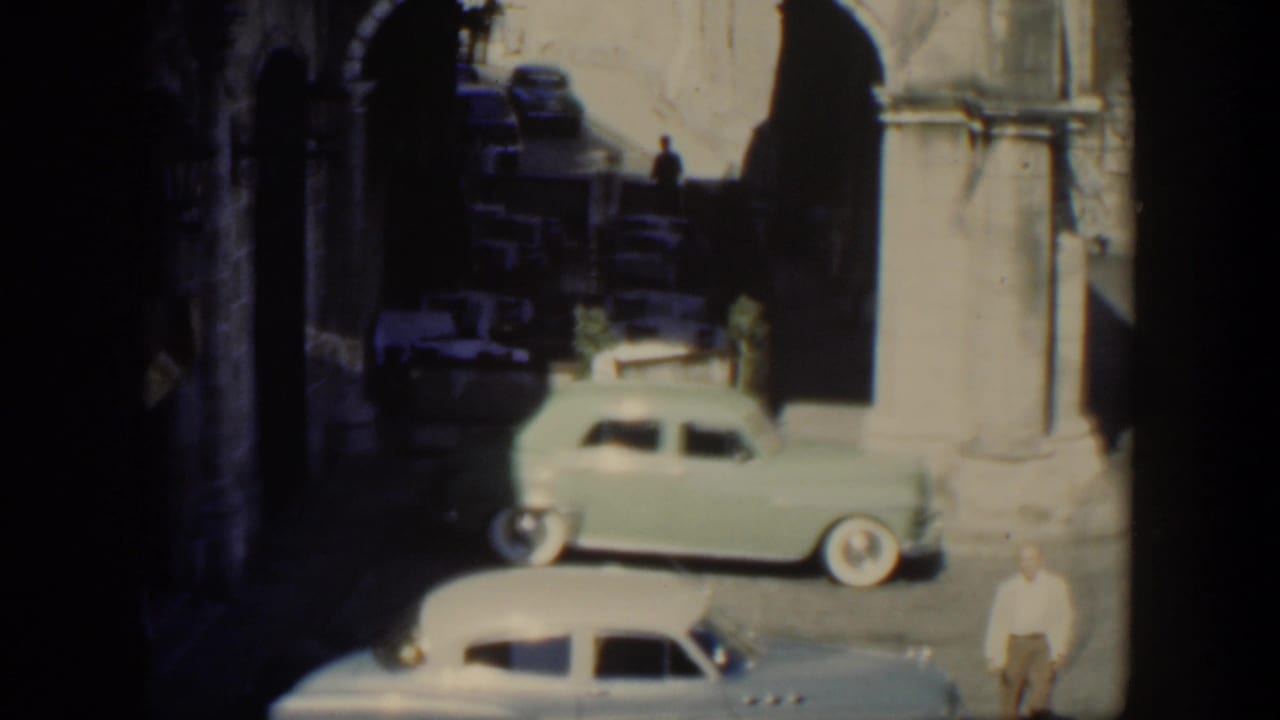 1951 Cuba home-movie frame showing classic cars on a narrow street beside weathered stone arches and deep shadow.