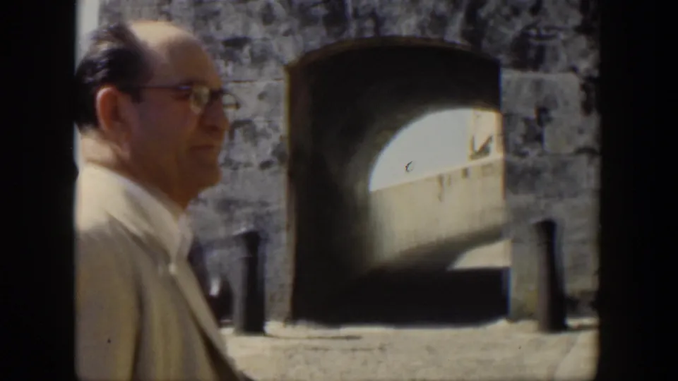 1951 Cuba frame of a man in a light-colored suit and glasses standing near a weathered stone arch opening.
