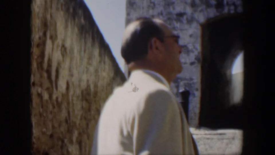 1951 Cuba home-movie view of a suited man in profile moving beside a tall stone wall toward an arched opening.