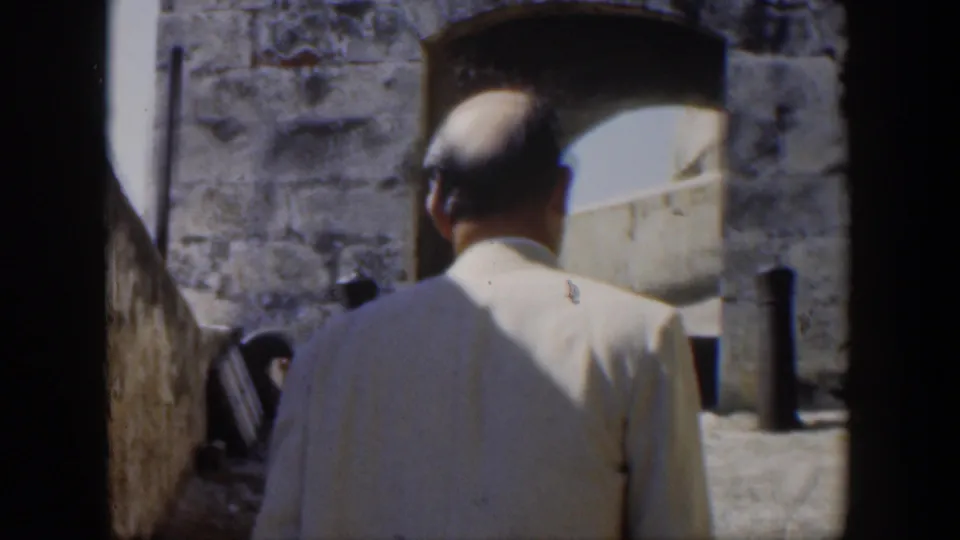 1951 Cuba frame showing the back of a man in a light suit as he faces a sunlit stone archway and masonry wall.