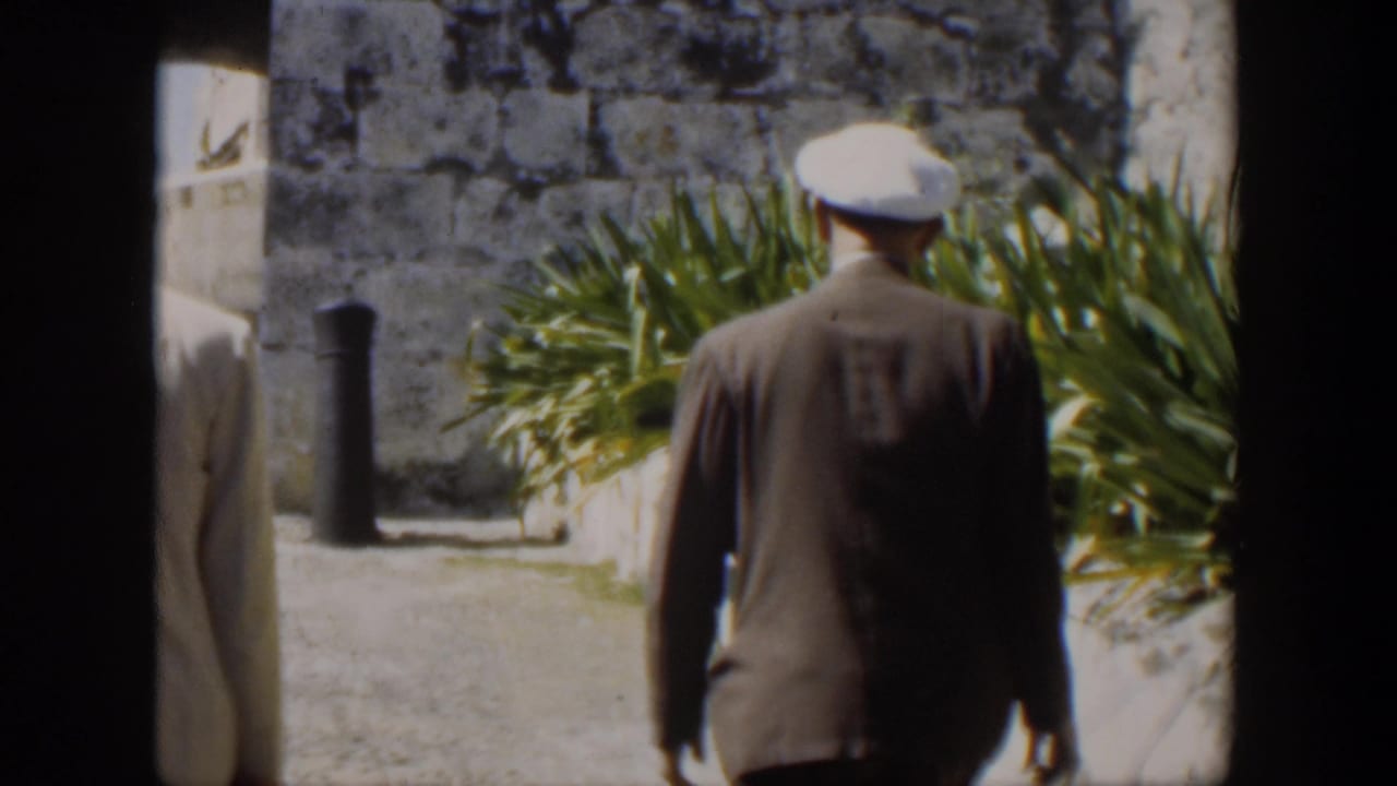 Man in dark suit and white cap walks beside plants and stone wall.