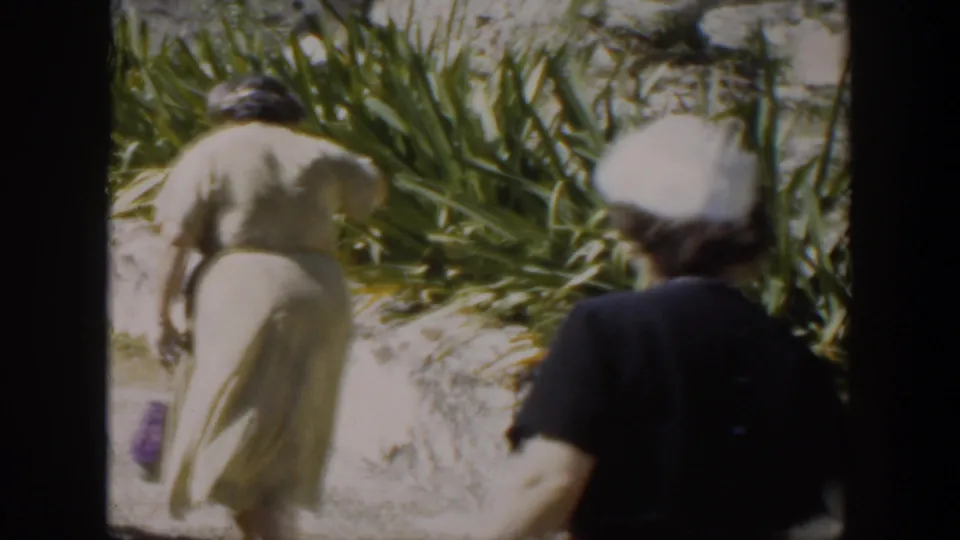 In the final frame, a woman in a light dress walks near plants while another adult in dark clothing crosses the foreground.