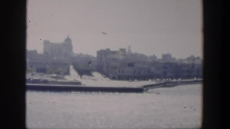 1951 Cuba 8mm frame of a hazy coastal city shoreline, with water in the foreground and low waterfront structures near the edge.