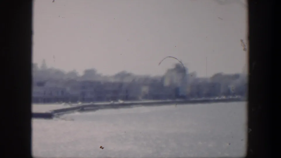 1951 Cuba 8mm frame of a hazy coastal city edge, with water in the foreground and indistinct buildings and trees along shore.