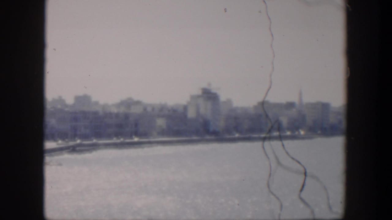 1951 Cuba home-movie frame showing a soft waterfront city view across blue water, with white buildings and film scratches.