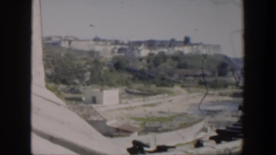 1951 Cuba 8mm frame showing a high view over stone fort walls, dark cannons, green trees, and pale buildings beyond.