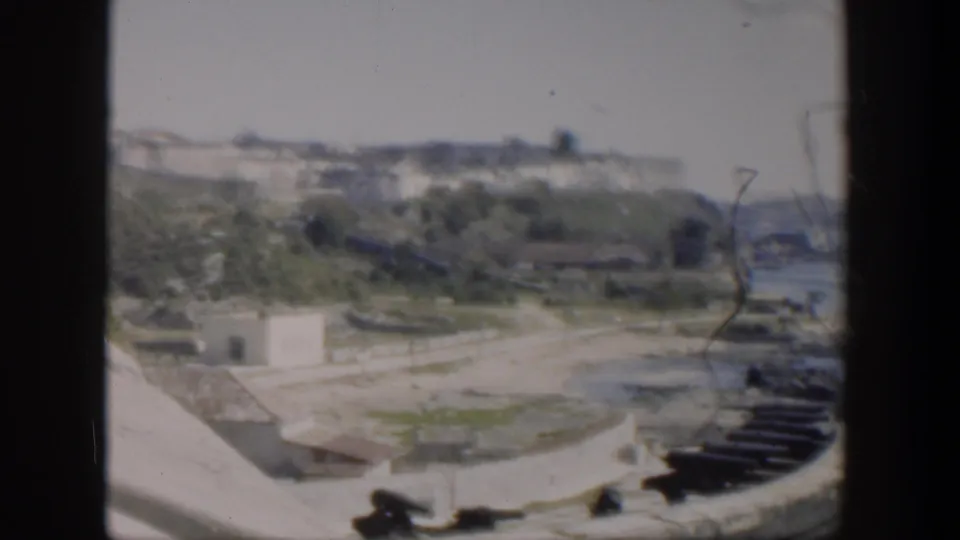 1951 Cuba home-movie frame with curved stone fortification, cannon positions, greenery, and distant white town buildings.