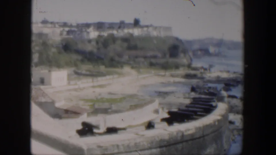 1951 Cuba 8mm view over waterfront stonework and cannons toward trees, pale buildings, and a hazy shoreline.