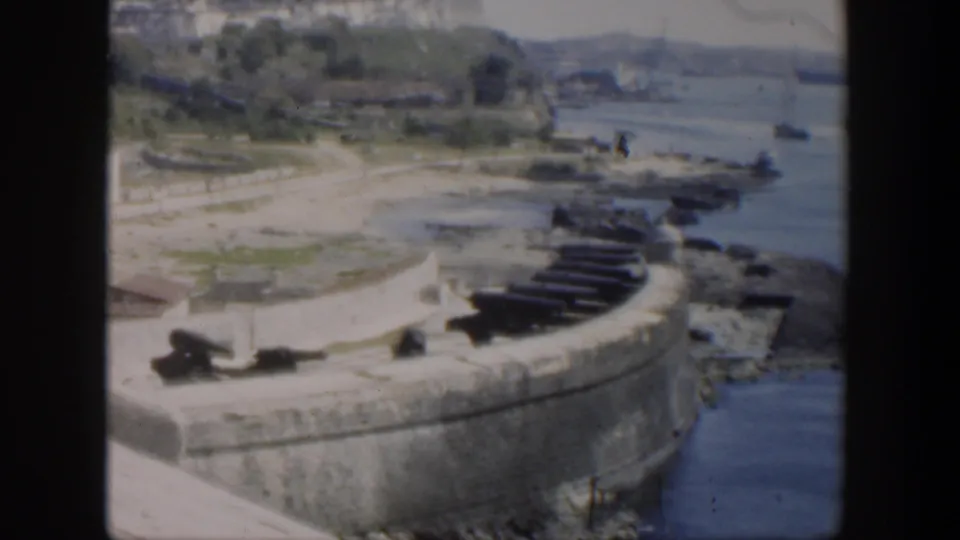 1951 Cuba home-movie frame showing the fort corner, cannon placements, shoreline water, green grounds, and distant townscape.