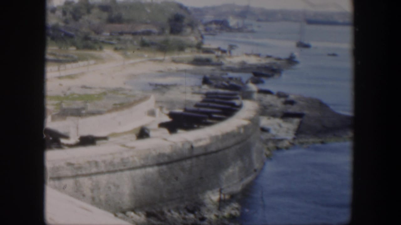 1951 Cuba home-movie frame showing cannon-lined stone ramparts above blue harbor water and rocky shore.