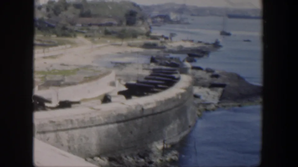 1951 Cuba home-movie frame showing cannon-lined stone ramparts above blue harbor water and rocky shore.