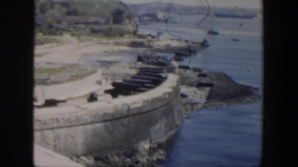 1951 Cuba view of a coastal fort rampart with cannons, shoreline rocks, calm water, and distant boats across the bay.