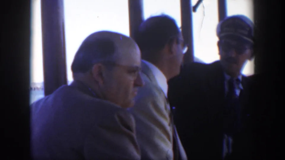 1951 Cuba home-movie frame showing three adult men in suits inside a vessel cabin, with window bars and bright exterior light behind them.
