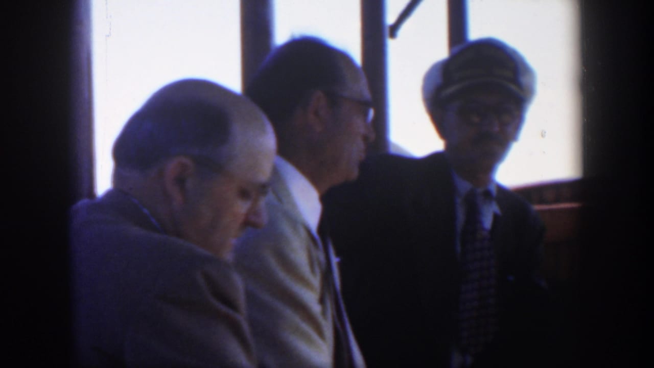 Older seated man and two other suited men, including a man in a peaked cap near the window.
