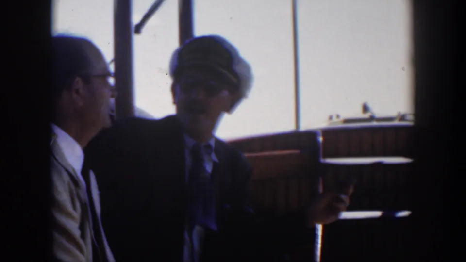 1951 Cuba home-movie frame of two suited men by vessel windows as the peaked-cap figure appears to gesture or hold a small item.