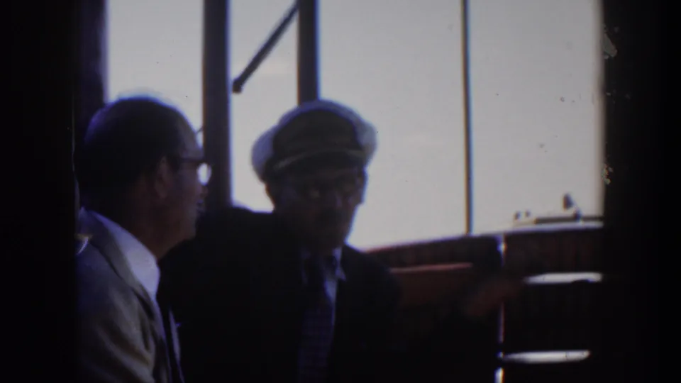 Dim 1951 Cuba ship-cabin frame with two adult men in suits, including a peaked-cap figure gesturing against bright window light.