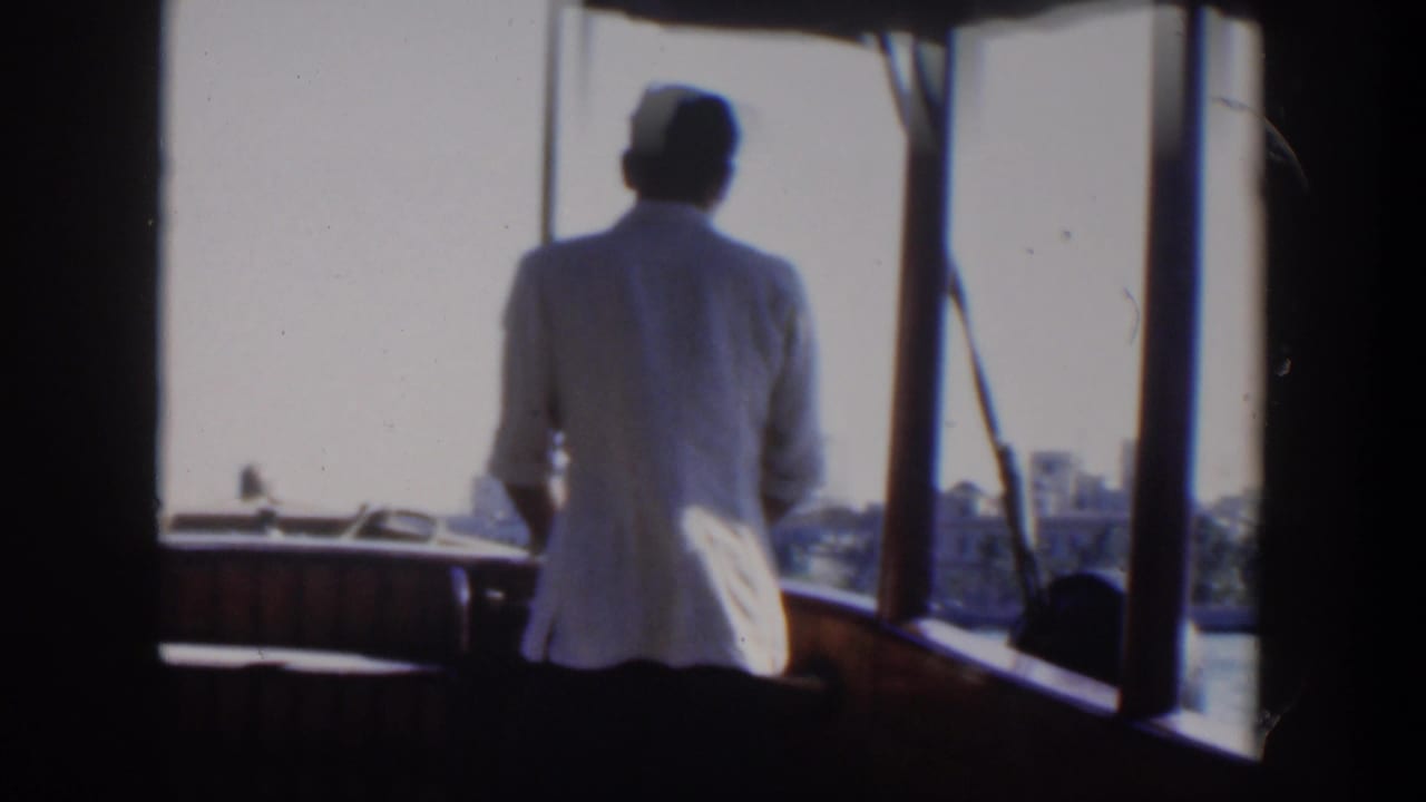 Man in a white jacket stands at a boat helm with water and shoreline ahead.